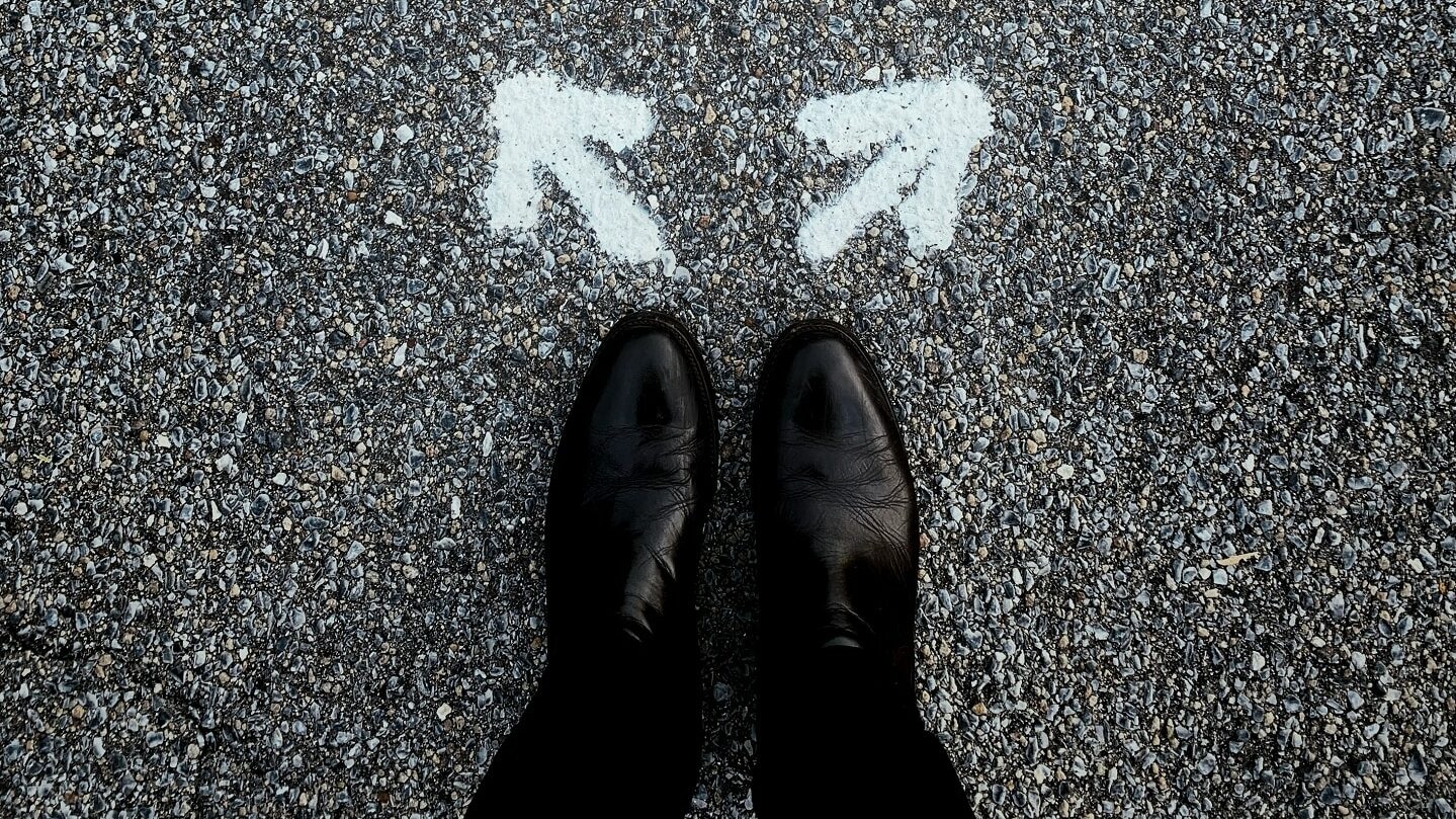 Chaussures noires sur le bitume, devant deux flèches blanches peintes qui partent l'une à gauche, l'autre à droite