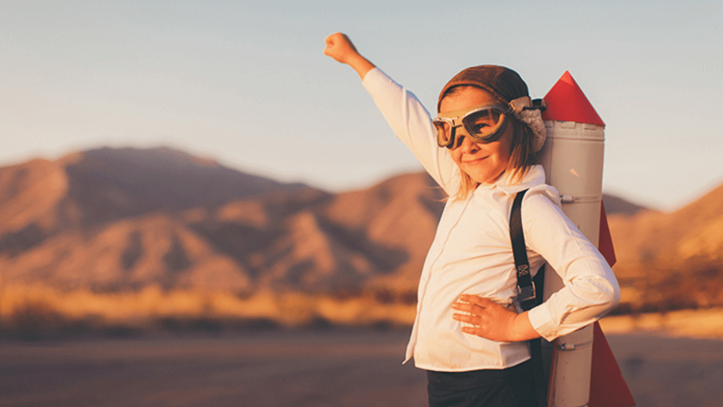 Enfant en chemise blanche et lunettes d’aviateur, une fusée en carton sur le dos, poing levé devant des montagnes au soleil couchant